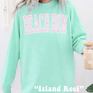 Może przedstawiać: Jasnozielona bluza z długim rękawem z napisem "Beach Bum" w r&oacute;żowych literach. Na dole bluzy nadrukowany jest napis "Island Reef" w kolorze białym.