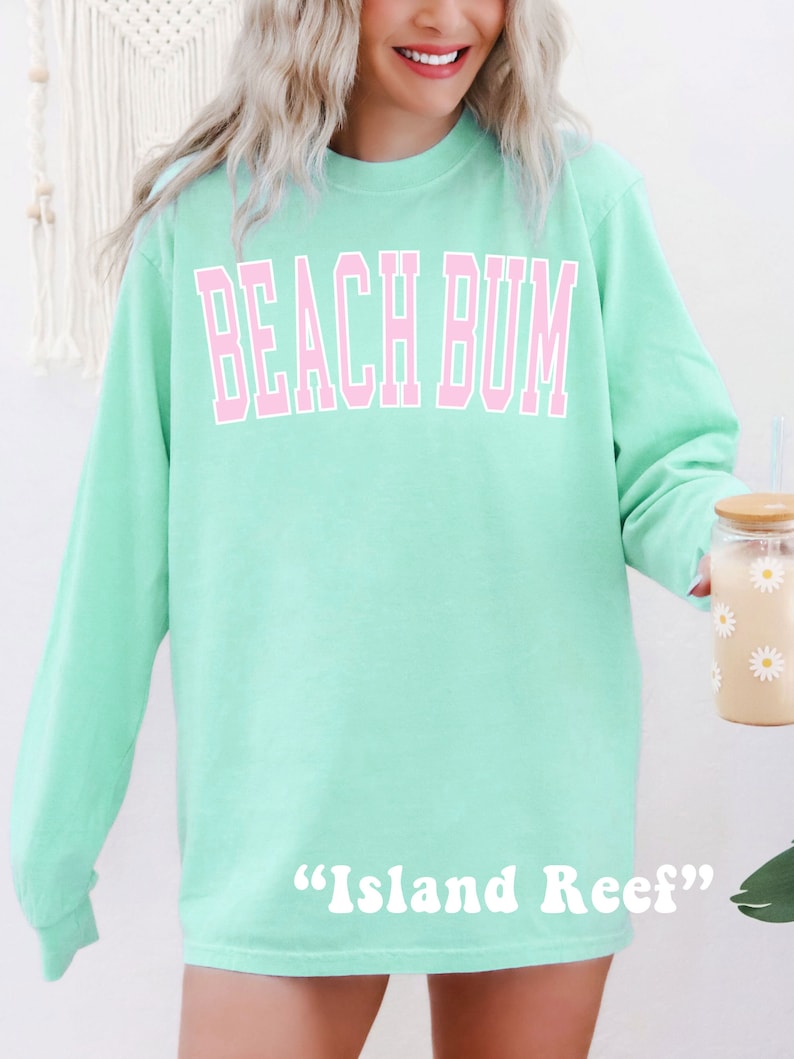 Może przedstawiać: Jasnozielona bluza z długim rękawem z napisem "Beach Bum" w r&oacute;żowych literach. Na dole bluzy nadrukowany jest napis "Island Reef" w kolorze białym.