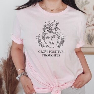 May include: A pink t-shirt with a black line drawing of a person with leaves growing out of their head. The text "GROW POSITIVE THOUGHTS" is printed below the drawing.