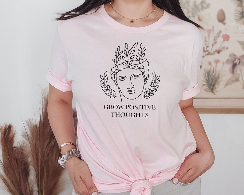 May include: A pink t-shirt with a black line drawing of a person with leaves growing out of their head. The text "GROW POSITIVE THOUGHTS" is printed below the drawing.