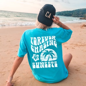 May include: A turquoise blue t-shirt with a white graphic that says "Forever Chasing Sunsets" with a palm tree and hibiscus flower design. The t-shirt is on a sandy beach with a person wearing a black baseball cap that says "CA" in gold letters.