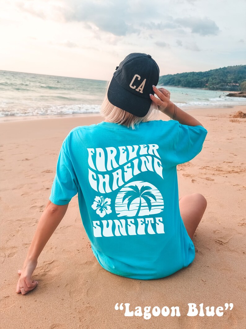May include: A turquoise blue t-shirt with a white graphic that says "Forever Chasing Sunsets" with a palm tree and hibiscus flower design. The t-shirt is on a sandy beach with a person wearing a black baseball cap that says "CA" in gold letters.