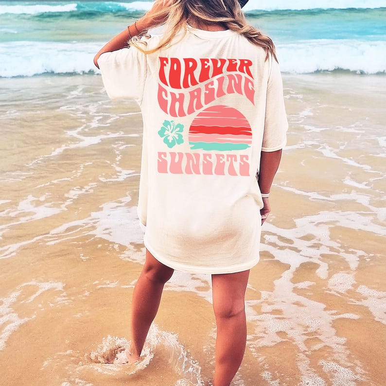 Pode incluir: Uma t-shirt de manga curta em tom off-white com um gr&aacute;fico nas costas com o texto "FOREVER CHASING SUNSETS" numa fonte vermelha ondulada. O design inclui um gr&aacute;fico de p&ocirc;r do sol estilizado em vermelho e turquesa e uma flor de hibisco.