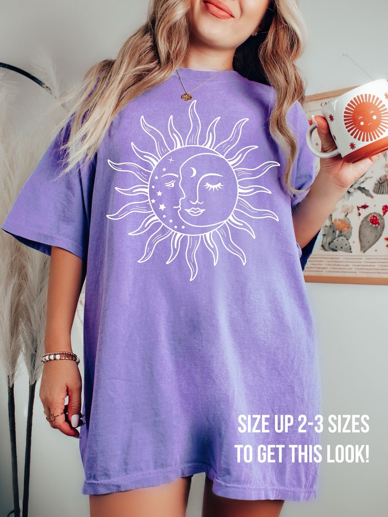 K&ouml;nnte beinhalten: Ein lilafarbenes Oversize-T-Shirt mit einem wei&szlig;en Sonnen- und Mondmotiv. Der Text "SIZE UP 2-3 SIZES TO GET THIS LOOK!" ist auf der Unterseite des Shirts aufgedruckt.