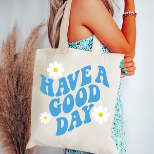 May include: A white canvas tote bag with blue and white daisy designs. The bag has blue text that reads "HAVE A GOOD DAY".