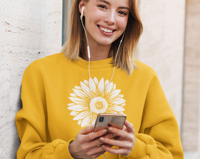 Pu&ograve; includere: Una felpa gialla con una grafica di girasole bianca. La felpa ha un collo rotondo e maniche lunghe.