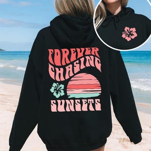 Pode incluir: Um moletom preto com capuz apresentando um gr&aacute;fico nas costas com o texto "FOREVER CHASING SUNSETS" em rosa, um p&ocirc;r do sol estilizado em rosa e turquesa, e uma flor de hibisco turquesa. A parte frontal inclui uma pequena flor de hibisco rosa.