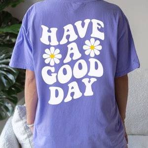 May include: A purple t-shirt with the text "HAVE A GOOD DAY" in white with daisy flowers. The text is in a retro font style. The t-shirt is labeled "Violet".