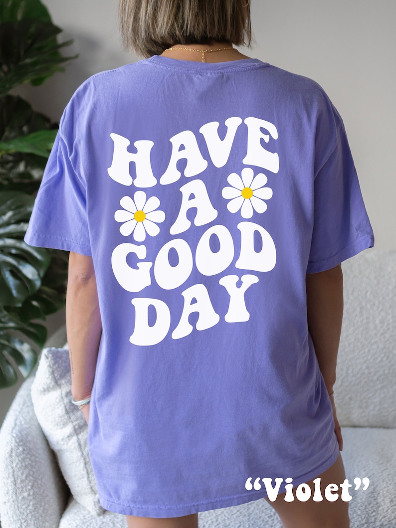 May include: A purple t-shirt with the text "HAVE A GOOD DAY" in white with daisy flowers. The text is in a retro font style. The t-shirt is labeled "Violet".