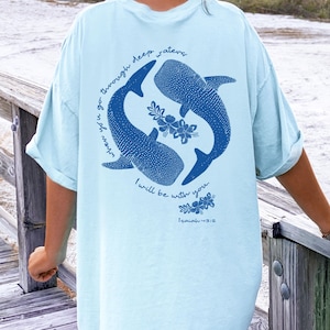 K&ouml;nnte beinhalten: Ein hellblaues T-Shirt mit einer dunkelblauen Grafik auf der R&uuml;ckseite. Das Design zeigt zwei Walhaie, die im Kreis schwimmen, mit floralen Akzenten und dem Text "when you go through deep waters I will be with you. Isaiah 43:2" in Blau.