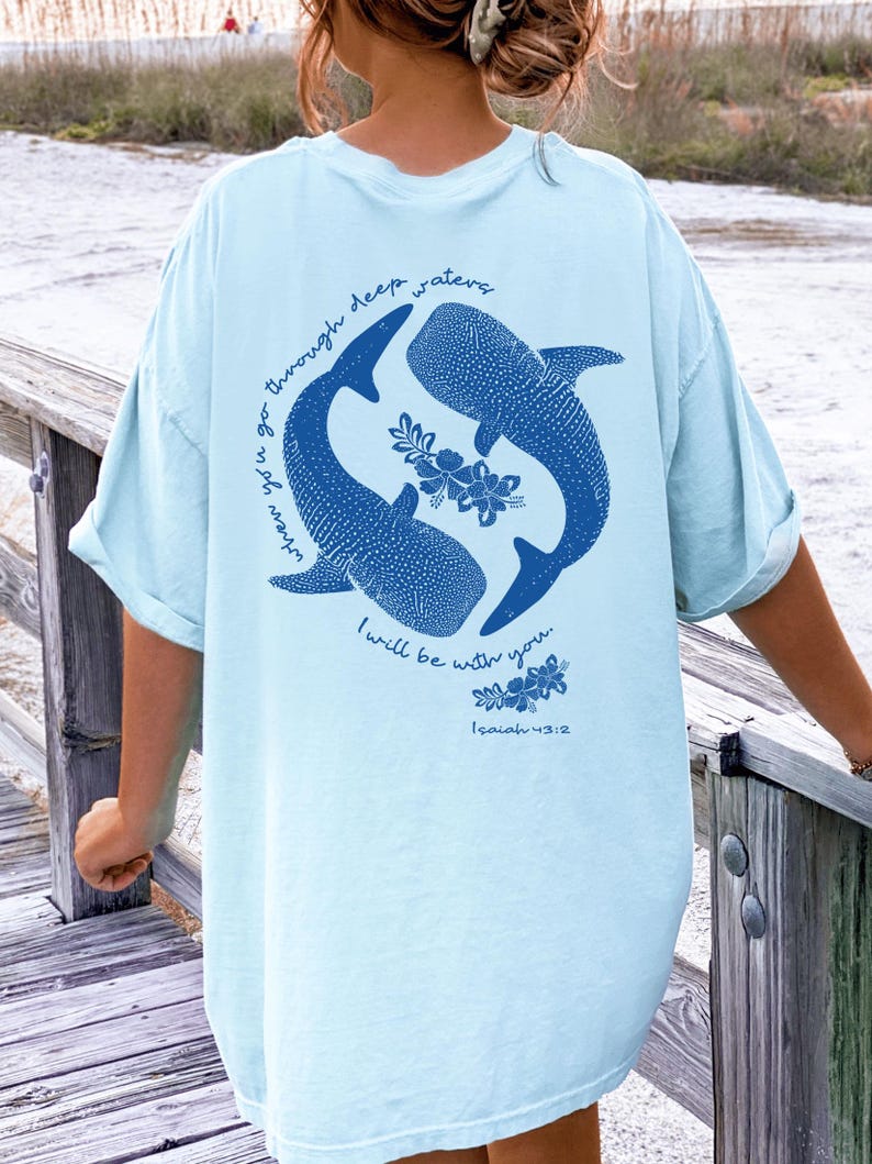 K&ouml;nnte beinhalten: Ein hellblaues T-Shirt mit einer dunkelblauen Grafik auf der R&uuml;ckseite. Das Design zeigt zwei Walhaie, die im Kreis schwimmen, mit floralen Akzenten und dem Text "when you go through deep waters I will be with you. Isaiah 43:2" in Blau.