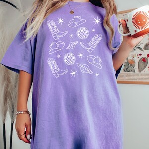 Puede incluir: Una camiseta de gran tama&ntilde;o de color morado con un dise&ntilde;o gr&aacute;fico blanco que presenta botas de vaquero, sombreros y bolas de discoteca. El texto "Violet" est&aacute; impreso debajo de la imagen.
