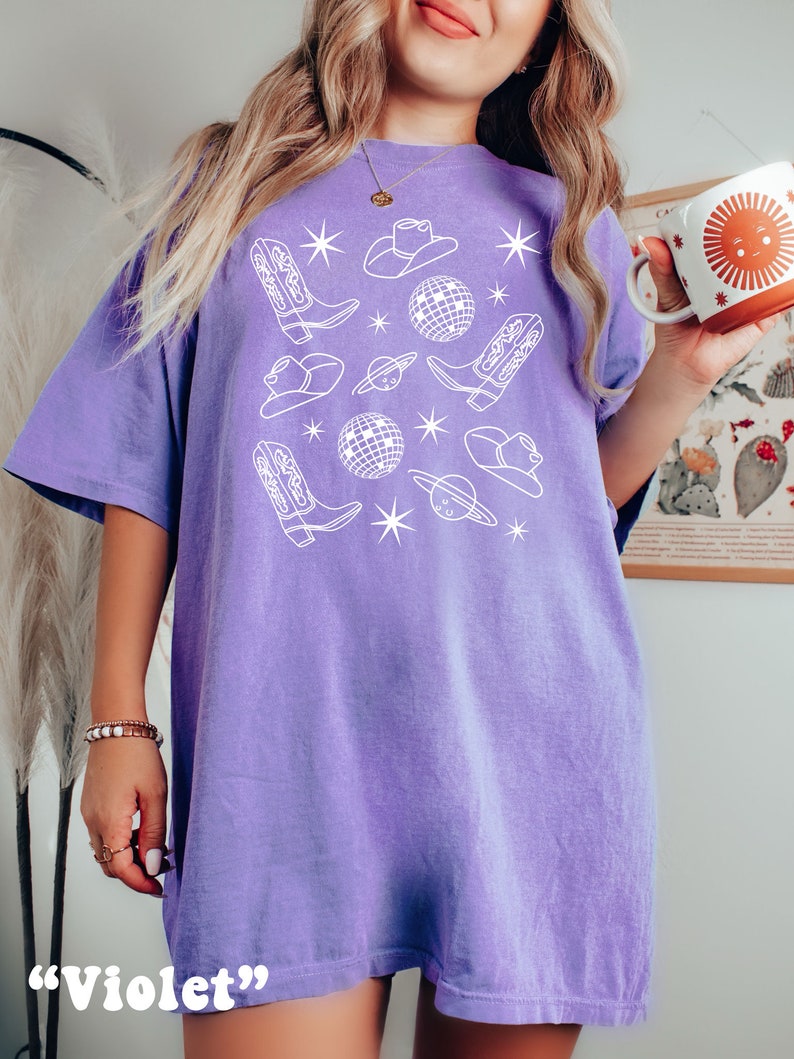Puede incluir: Una camiseta de gran tama&ntilde;o de color morado con un dise&ntilde;o gr&aacute;fico blanco que presenta botas de vaquero, sombreros y bolas de discoteca. El texto "Violet" est&aacute; impreso debajo de la imagen.