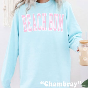 Może przedstawiać: Jas sportowy w jasnoniebieskim kolorze z napisem "Beach Bum" w r&oacute;żowych literach. Na dole znajduje się metka "Chambray".