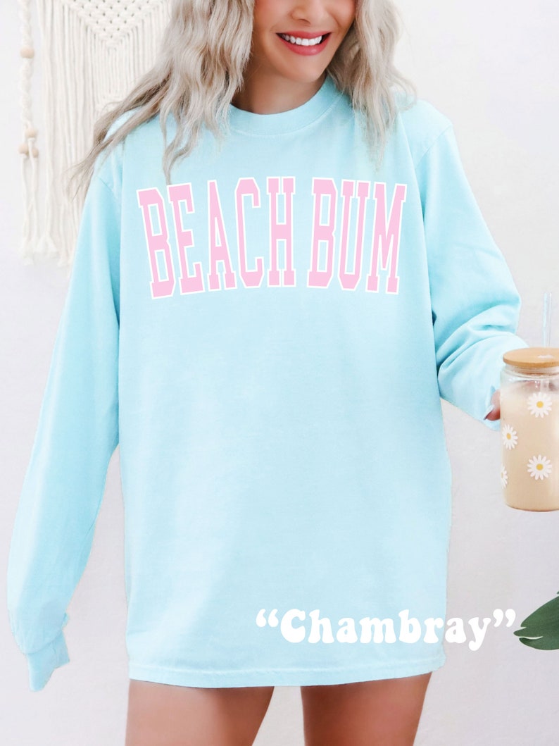 Może przedstawiać: Jas sportowy w jasnoniebieskim kolorze z napisem "Beach Bum" w r&oacute;żowych literach. Na dole znajduje się metka "Chambray".
