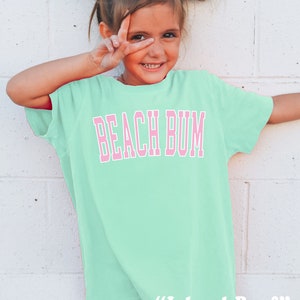 May include: A light green t-shirt with the words "BEACH BUM" in pink block letters. The shirt is worn by a young person with white bows in their hair, making a peace sign with their fingers. The text "Island Reef" is printed at the bottom.