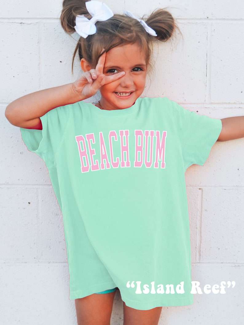 May include: A light green t-shirt with the words "BEACH BUM" in pink block letters. The shirt is worn by a young person with white bows in their hair, making a peace sign with their fingers. The text "Island Reef" is printed at the bottom.