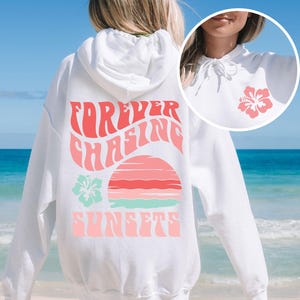 Pode incluir: Uma camisola com capuz branca com um gr&aacute;fico rosa e turquesa nas costas com o texto "FOREVER CHASING SUNSETS" sobre um sol estilizado e uma flor de hibisco. Um gr&aacute;fico de hibisco mais pequeno est&aacute; impresso na zona do peito.