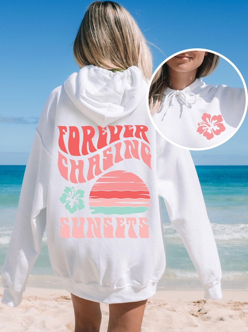Pode incluir: Uma camisola com capuz branca com um gr&aacute;fico rosa e turquesa nas costas com o texto "FOREVER CHASING SUNSETS" sobre um sol estilizado e uma flor de hibisco. Um gr&aacute;fico de hibisco mais pequeno est&aacute; impresso na zona do peito.