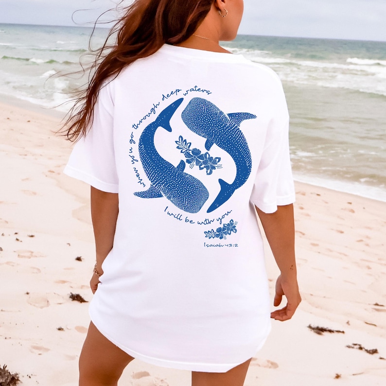 K&ouml;nnte beinhalten: Ein wei&szlig;es Kurzarm-T-Shirt mit einer blauen Grafik auf der R&uuml;ckseite. Das Design zeigt zwei Walhaie in kreisf&ouml;rmiger Anordnung mit floralen Akzenten. Der Text lautet: "when you go through deep waters I will be with you. Isaiah 43:2."