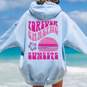Pode incluir: Um moletom com capuz azul-claro apresentando um gr&aacute;fico nas costas com o texto "FOREVER CHASING SUNSETS" em rosa. O design inclui um gr&aacute;fico estilizado de p&ocirc;r do sol em rosa e roxo e um &iacute;cone de flor de hibisco roxa no lado esquerdo.