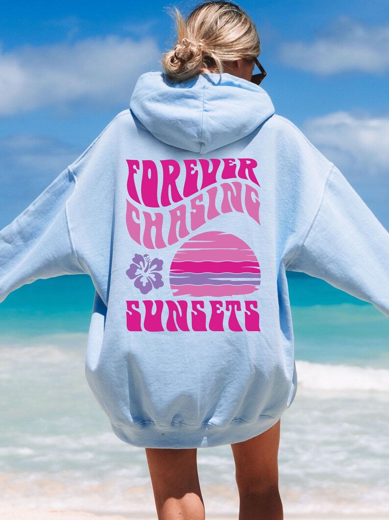 Pode incluir: Um moletom com capuz azul-claro apresentando um gr&aacute;fico nas costas com o texto "FOREVER CHASING SUNSETS" em rosa. O design inclui um gr&aacute;fico estilizado de p&ocirc;r do sol em rosa e roxo e um &iacute;cone de flor de hibisco roxa no lado esquerdo.
