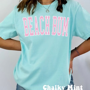 May include: A light blue t-shirt with the text "Beach Bum" in pink lettering. The shirt is being modeled by a person wearing denim shorts. The text "Chalky Mint" is written in white at the bottom of the image.
