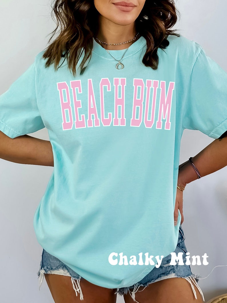 May include: A light blue t-shirt with the text "Beach Bum" in pink lettering. The shirt is being modeled by a person wearing denim shorts. The text "Chalky Mint" is written in white at the bottom of the image.