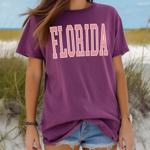 Op de afbeelding: Een paars T-shirt met het woord "FLORIDA" bedrukt in roze letters met witte contouren. Het shirt is oversized en heeft een relaxte pasvorm. Het model draagt het shirt met een spijkerbroekje en staat op een zandstrand.