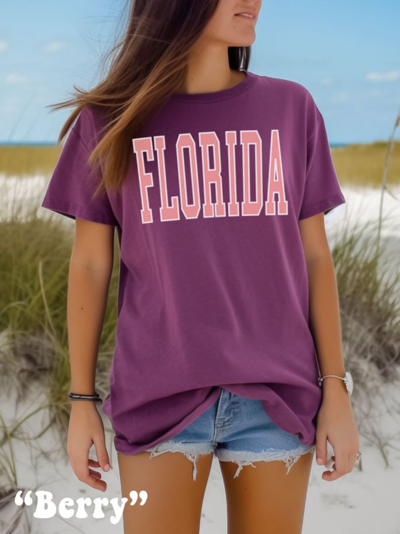 Op de afbeelding: Een paars T-shirt met het woord "FLORIDA" bedrukt in roze letters met witte contouren. Het shirt is oversized en heeft een relaxte pasvorm. Het model draagt het shirt met een spijkerbroekje en staat op een zandstrand.