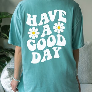 May include: A turquoise blue t-shirt with a white graphic that says "Have a Good Day" with two white daisies on either side of the "a".