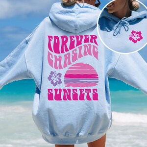 Pode incluir: Um moletom com capuz azul claro apresentando um gr&aacute;fico grande nas costas com o texto "FOREVER CHASING SUNSETS" em rosa, acompanhado por uma ilustra&ccedil;&atilde;o de p&ocirc;r do sol rosa e roxo e uma flor de hibisco. Um pequeno hibisco est&aacute; na frente.