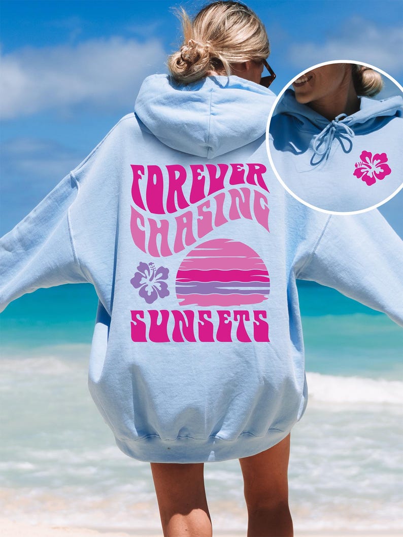 Pode incluir: Um moletom com capuz azul claro apresentando um gr&aacute;fico grande nas costas com o texto "FOREVER CHASING SUNSETS" em rosa, acompanhado por uma ilustra&ccedil;&atilde;o de p&ocirc;r do sol rosa e roxo e uma flor de hibisco. Um pequeno hibisco est&aacute; na frente.