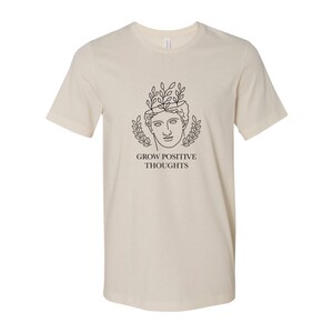 May include: A beige t-shirt with a black line drawing of a person's head with leaves growing out of it. The text "GROW POSITIVE THOUGHTS" is printed below the drawing.