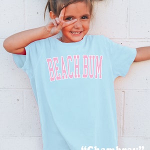 May include: A light blue t-shirt with the words "BEACH BUM" in pink block letters. The shirt is worn by a young person with white bows in their hair. The word "Chambray" is written in white at the bottom of the shirt.