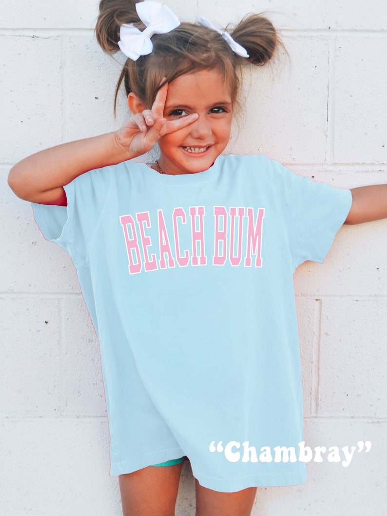 May include: A light blue t-shirt with the words "BEACH BUM" in pink block letters. The shirt is worn by a young person with white bows in their hair. The word "Chambray" is written in white at the bottom of the shirt.