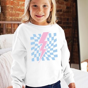 Puede incluir: Sudadera blanca con un gr&aacute;fico de rayo rosa sobre un fondo a cuadros azul y blanco.
