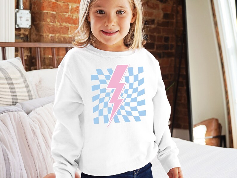 Puede incluir: Sudadera blanca con un gr&aacute;fico de rayo rosa sobre un fondo a cuadros azul y blanco.