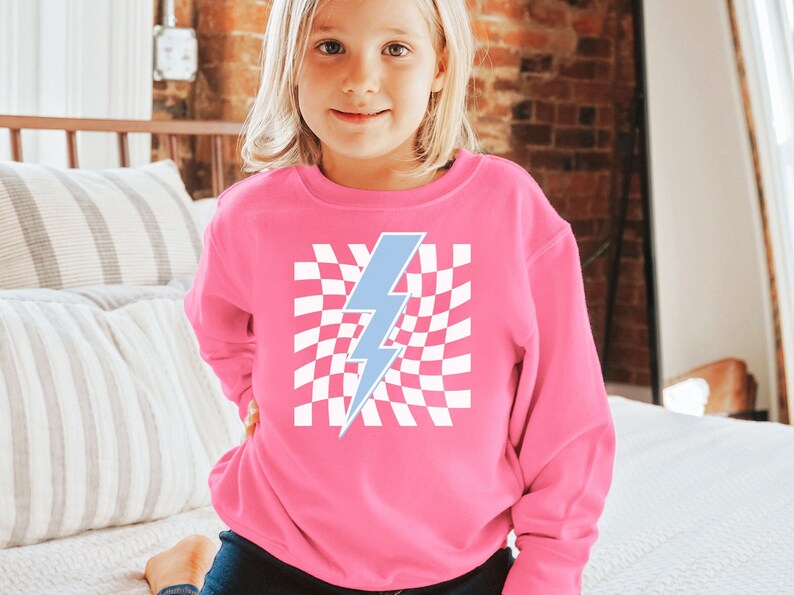 Puede incluir: Una sudadera rosa con un gr&aacute;fico de rayo azul sobre un fondo a cuadros blancos.