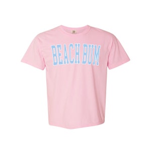 Pu&ograve; includere: Una maglietta rosa con la scritta "BEACH BUM" in azzurro.