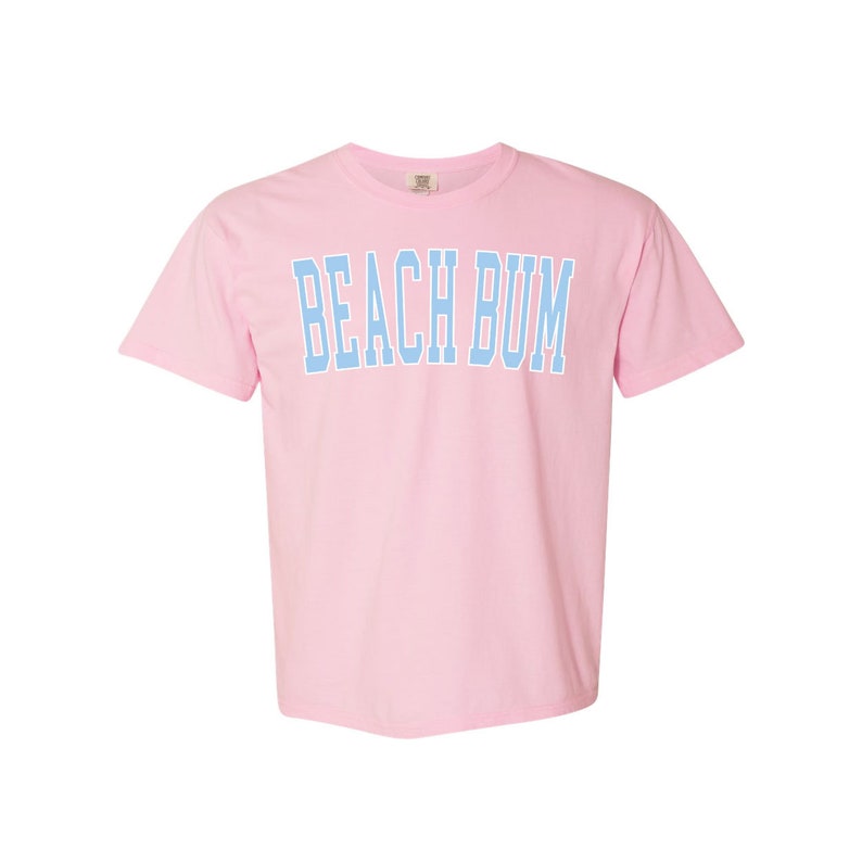 Pu&ograve; includere: Una maglietta rosa con la scritta "BEACH BUM" in azzurro.