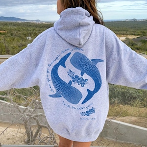 K&ouml;nnte beinhalten: Ein hellgrauer Kapuzenpullover mit einer blauen Grafik auf der R&uuml;ckseite. Das Design zeigt zwei Walhaie in kreisf&ouml;rmiger Anordnung, umgeben von floralen Akzenten und dem Text: When you go through deep waters I will be with you. Isaiah 43:2.
