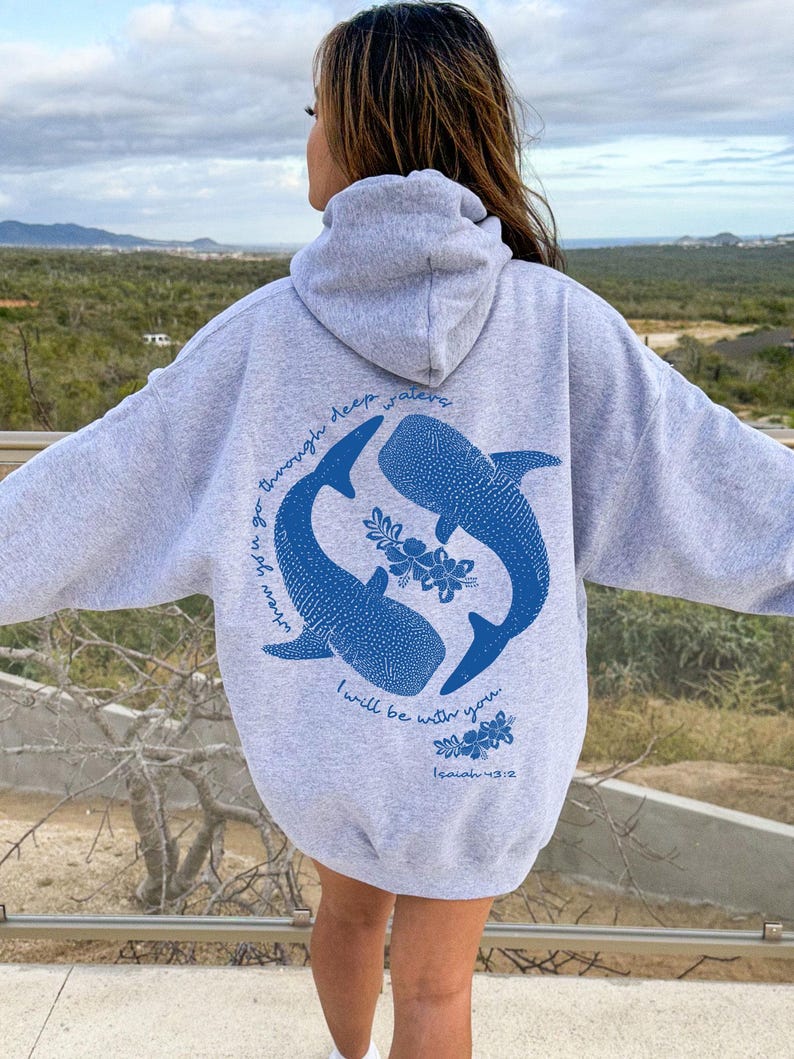 K&ouml;nnte beinhalten: Ein hellgrauer Kapuzenpullover mit einer blauen Grafik auf der R&uuml;ckseite. Das Design zeigt zwei Walhaie in kreisf&ouml;rmiger Anordnung, umgeben von floralen Akzenten und dem Text: When you go through deep waters I will be with you. Isaiah 43:2.