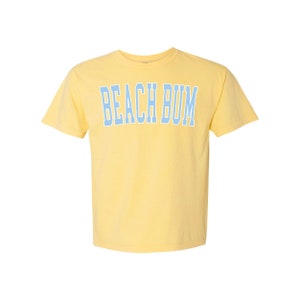 Pu&ograve; includere: Una maglietta gialla a maniche corte con il testo "BEACH BUM" in blu.