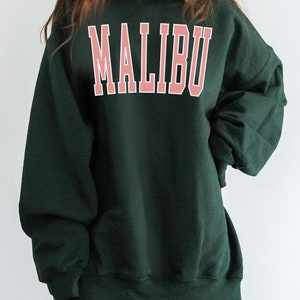 K&ouml;nnte beinhalten: Ein dunkelgr&uuml;ner Oversize-Sweatshirt mit dem Wort "MALIBU" in Pink auf der Vorderseite.