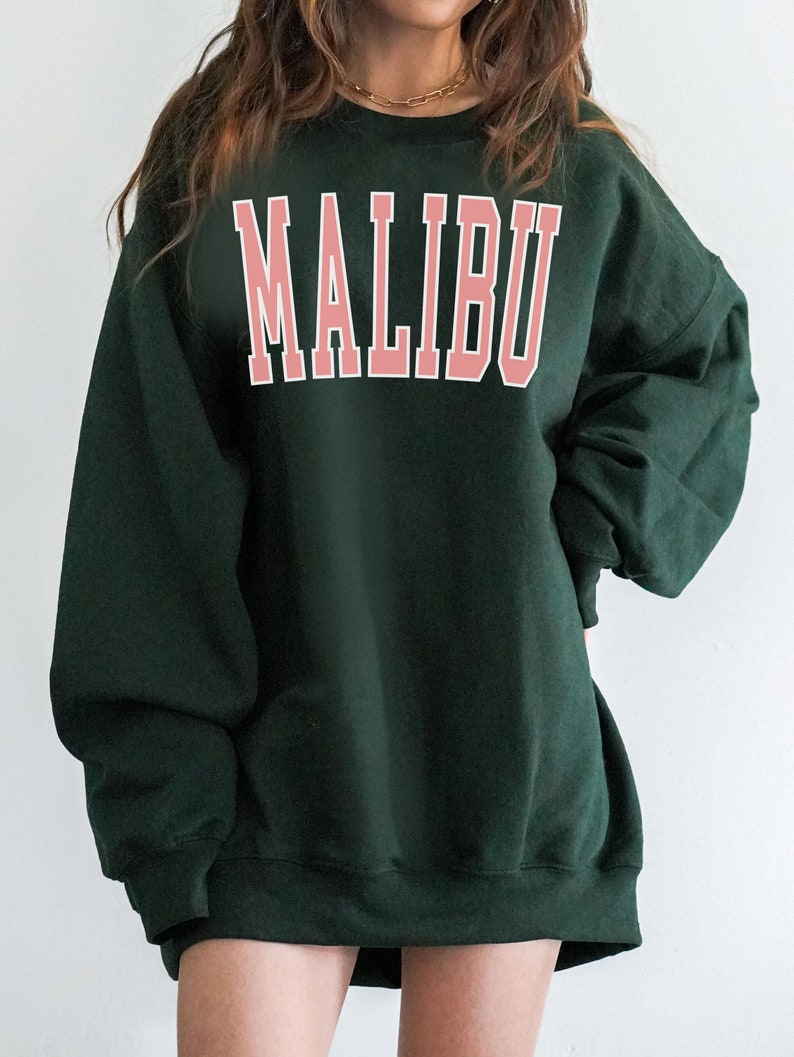 K&ouml;nnte beinhalten: Ein dunkelgr&uuml;ner Oversize-Sweatshirt mit dem Wort "MALIBU" in Pink auf der Vorderseite.