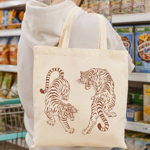 May include: A white tote bag with brown handles features a graphic design of two tigers. The tigers are facing opposite directions and have a stylized, traditional Japanese design. The bag is made of a natural fabric and has a simple, elegant look.
