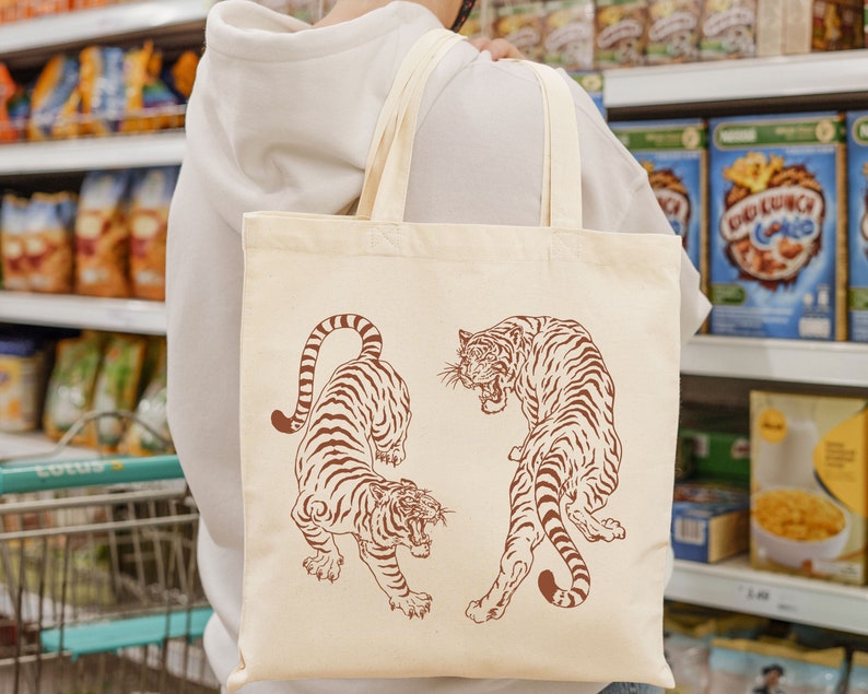 May include: A white tote bag with brown handles features a graphic design of two tigers. The tigers are facing opposite directions and have a stylized, traditional Japanese design. The bag is made of a natural fabric and has a simple, elegant look.