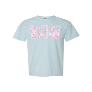 May include: Light blue t-shirt with the text "BEACH BUM" in pink block letters. The shirt has short sleeves and a crew neck. The text is outlined in white.
