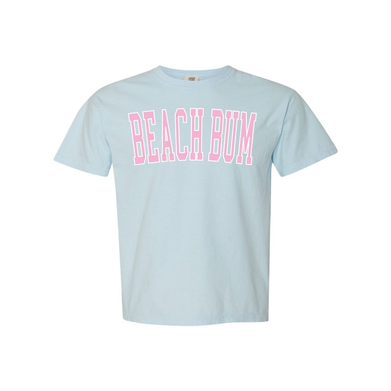 May include: Light blue t-shirt with the text "BEACH BUM" in pink block letters. The shirt has short sleeves and a crew neck. The text is outlined in white.
